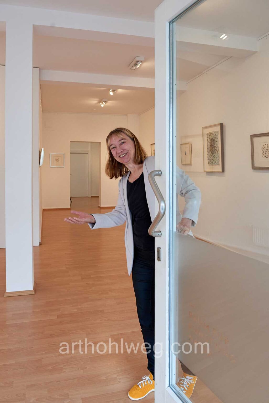 Magdalena Hohlweg öffnet die Glastür zu einer Galerie und weist in den Ausstellungsraum mit ihren gerahmten Arbeiten.